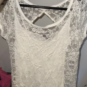 Tröja/blus - Säljer en vit tröja med blommig spets och snygg öppen ryggdetalj. Blusen har korta ärmar och ett tunt innerfoder som gör den bekväm att bära. Perfekt för dig som gillar romantisk och feminin stil med extra detaljer.