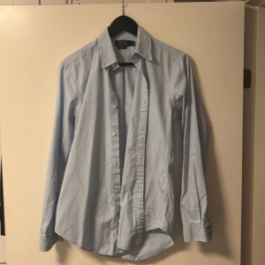Ljusblå skjorta Polo Ralph Lauren S - Snygg ljusblå skjorta från Polo Ralph Lauren i slim fit-modell. Skjortan har klassisk krage, långa ärmar med manschett och knappar framtill. Tillverkad i mjuk bomull som känns skön mot huden. Perfekt för dig som gillar stilrena och fräscha plagg.