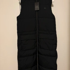 Svart lång dunväst från Equine XS - Snygg svart lång dunväst från Equine i storlek XS. Västen har huva, två stora fickor framtill och dragkedja hela vägen. Perfekt för lager-på-lager och har en stilren, quiltad look som funkar till många outfits. 