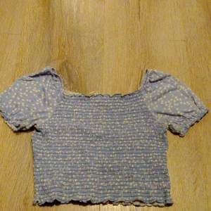 Blommig blå croppad topp Kappahl - Söt croppad topp från Kappahl i ljusblått med små vita blommor. Toppen har kort puffärm och smockad kropp som ger en tight och stretchig passform. Perfekt för dig som gillar romantisk och trendig stil.