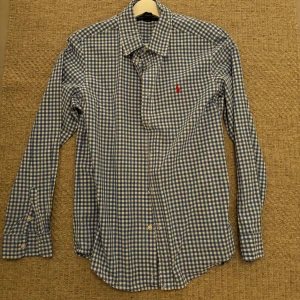 Blårutig skjorta från Ralph Lauren - Klassisk blårutig skjorta från Ralph Lauren med lång ärm och knappar framtill. Skjortan har en vit och blå rutig design och ikonisk röd broderad logga på bröstet. Tillverkad i mjuk bomull och perfekt för en clean och stilren look. Storlek S. Tveka inte att höra av sig vid frågor!