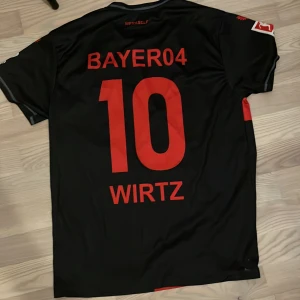 Bayer Leverkusen Wirtz matchtröja 10/10 skick - Säljer en svart Bayer Leverkusen fotbollströja från Castore med röd korsdesign på framsidan, tryck BAYER04 och nummer 10 Wirtz på ryggen. Tröjan har klubbmärke broderat på bröstet och sponsortryck. Klassisk rund hals och korta ärmar. Perfekt för fans!