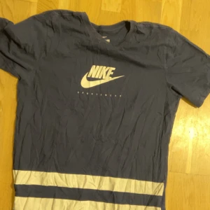 Blå Nike t-shirt med logga - Snygg blå t-shirt från Nike med vit logga och text på bröstet. T-shirten har korta ärmar och två vita ränder längst ner. Skön passform och tillverkad i mjuk bomull, perfekt för en sportig look.