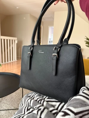 Svart handväska från Rizzo - Stilren svart handväska från Rizzo med två rejäla handtag och silverfärgade detaljer. Väskan har flera fack med dragkedja och en praktisk ficka på baksidan. Materialet är skinnimitation med diskret struktur. Perfekt för dig som gillar ordning och vill ha plats för allt viktigt. Man får plats med en skoladata.