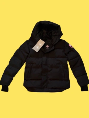 Svart pufferjacka från Canada Goose - Svart pufferjacka från Canada Goose med huva och klassisk logga på ärmen. Jackan har dragkedja framtill, två stora fickor och ribbade muddar vid ärmslut. Perfekt för kalla dagar och har en clean, modern look som funkar till streetwear.