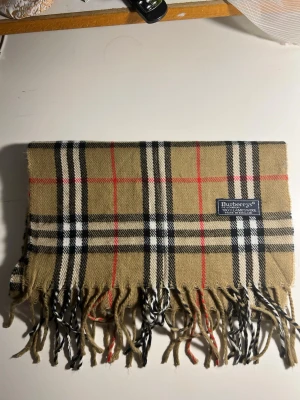 Rutig beige Burberrys halsduk i ull - Säljer en klassisk rutig halsduk från Burberrys i beige, svart, vit och röd. Västen är tillverkad i 100% lammull och har fransar längs nederkanten. Perfekt för dig som gillar tidlös design och vill ha något som sticker ut. (2A)
