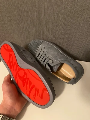 Grå Christian Louboutin sneakers med nitar - Säljer ett par grå sneakers från Christian Louboutin med coola nitar på tån och klassisk röd sula med logga. Skorna är gjorda i mocka och har snörning samt rund tå. Perfekt för dig som vill sticka ut med lyxig streetstyle.