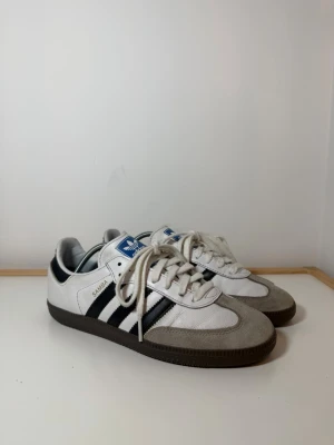 Adidas Samba sneakers vit och svart - Adidas Samba sneakers med klassiska svarta ränder på sidorna, vit ovandel i skinn och grå mockadetaljer vid tån. Skorna har snörning, platt sula och rund tå. Blå Adidas-logga på plösen. Perfekt för dig som gillar retrostil och sportig vibe.