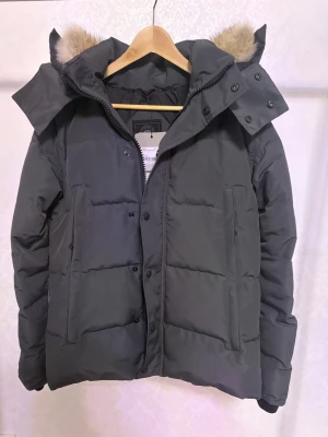 Canada Goose Wyndham Jacka - Hej! Säljer en grå Canada Goose Jacka modell: Wyndham med black label. pälskantad huva och klassisk logga på ärmen. Jackan har en pälskantad huva med dragkedja och knappar framtill, och en varm, vadderad insida. Perfekt för kalla dagar och riktigt snygg streetstil 😊❄️