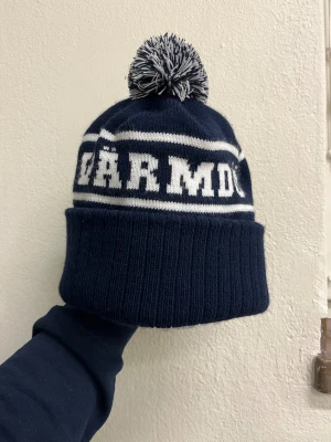 Mörkblå mössa med tofs  - Säljer en mörkblå stickad mössa med vit text och randdetaljer. Mössan har en stor tofs upptill i blått och vitt och bred uppvikt kant. Perfekt accessoar för kalla dagar och snygg till vinterlooken.