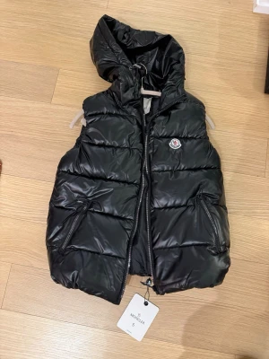 Svart dunväst från Moncler med huva - Säljer en svart dunväst från Moncler med huva och två praktiska fickor med dragkedja. Västen har quiltad design och ett snyggt Moncler-märke på bröstet. Perfekt för lager-på-lager och riktigt chill stil.