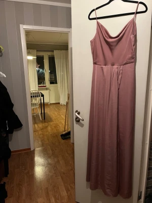 Rosa långklänning från Bubbleroom - Säljer en elegant ljusrosa långklänning från Bubbleroom med smala axelband och snygg draperad urringning. Klänningen har dekorativa knappar baktill och är tillverkad i ett mjukt, glansigt polyestertyg som faller fint. Perfekt för dig som vill ha en stilren look.