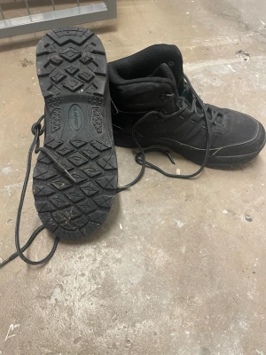 Svarta kängor från Hi-Tec - Svarta kängor från Hi-Tec med grov mönstrad sula och snörning hela vägen upp. Skorna har en robust och sportig design med vadderad krage och tålig ovandel i syntetmaterial. Perfekta för utomhusaktiviteter och äventyr.