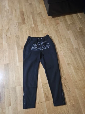 Svarta mjukisbyxor från Trapstar S - Svarta mjukisbyxor från Trapstar med broderad logga och text framtill. Byxorna har snörning i midjan och dragkedjor vid bensluten. Tillverkade i mjukt bomullsmaterial för skön känsla. Perfekta för chill eller streetwear.