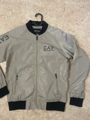 Beige bomberjacka från EA7 Emporio Armani - Snygg beige bomberjacka från EA7 Emporio Armani med svarta muddar och krage. Jackan har dragkedja framtill, två fickor med zip och tryckt logga på bröstet och ena ärmen. Perfekt för dig som vill ha en clean och stilren look.