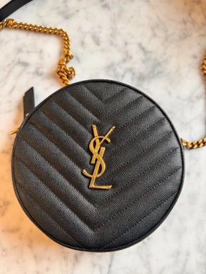 Svart rund axelväska från YSL - Säljer en rund svart axelväska från YSL i kalvskinn med quiltad chevronmönster och guldfärgad kedja. Väskan har YSL-logga i metall framtill, dragkedja upptill och ett huvudfack med en platt innerficka. Perfekt storlek för det viktigaste.