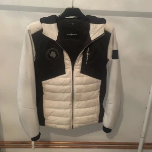 Vit och svart pufferjacka Sail Racing - Snygg pufferjacka från Sail Racing i vit och svart med broderad Sail Antarctica Expedition-logga på bröstet. Jackan har huva, dragkedja och quiltad front. Perfekt för dig som gillar sportig stil och vill ha en jacka som sticker ut.