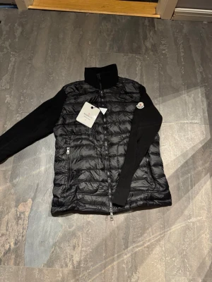 Moncler cardigan - Säljer en svart pufferjacka från Moncler med quiltad kropp och stickade ärmar. Jackan har hög krage, dragkedja framtill och två fickor med dragkedja. Klassisk Moncler-logga på ärmen och coolt serietryck på insidan. Perfekt för dig som vill ha både stil och komfort.