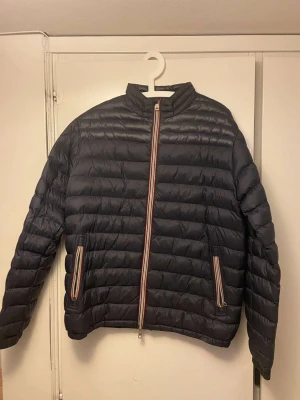 Mörkblå Moncler pufferjacka - Snygg mörkblå pufferjacka från Moncler med horisontella quiltade paneler och ståkrage. Jackan har dragkedja med röd, vit och blå randiga detaljer samt matchande dragkedjor på fickorna. Perfekt för kyliga dagar och har en clean look.