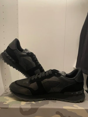 Valentino Garavani svarta sneakers - Säljer ett par svarta Valentino Garavani sneakers med snyggt kamouflagemönster och nitar på sulan. Skorna har rund tå, snörning och är tillverkade i en mix av skinn och syntetmaterial. Perfekta för dig som gillar exklusiv streetstyle.