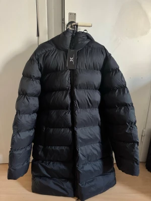 Svart puffer parka från Cross Sportswear - Svart puffer parka från Cross Sportswear i storlek M. Jackan har en hög krage, huva och tydliga quiltade sektioner. Materialet är syntet och dun, med dragkedja framtill och en diskret logga på ärmen. Perfekt för kalla dagar när du vill hålla dig varm och se chill ut.