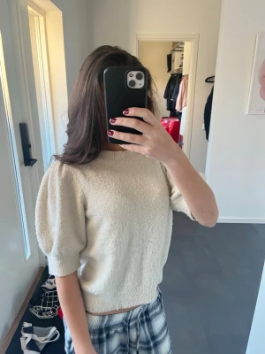Beige stickad topp med puffärm från vera moda  - Säljer en beige stickad topp med puffiga korta ärmar och rund hals. Toppen är croppad och har en mjuk, lite fluffig struktur som gör den extra mysig. Perfekt att styla med jeans eller kjol för en chill look.