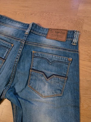 Blå Diesel jeans  - Säljer ett par klassiska blå jeans från Diesel med snygga slitningar och raka ben. Jeansen har fem fickor, tydliga kontrastsömmar och en läderpatch med Diesel-logga bak. Passar dig som gillar en avslappnad och tidlös stil.