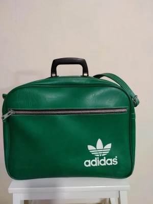 Vintage Adidas väska med axelrem  - Vintage Adidas väska från 70-talet. Har ett runt tryckmärke på utsidan och bruksslitage på in- och utsidan. Material: PVC