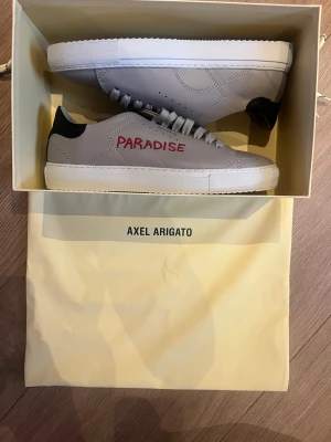 Axel Arigato Paradise sneakers grå - Snygga grå sneakers från Axel Arigato med vit sula och svart häl. Skorna har snörning och texten 'PARADISE' broderad i rött på sidan. Storlek 39                                                                               Skriv för fler bilder eller för att diskutera pris 