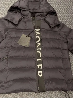 Svart pufferjacka från Moncler - Svart pufferjacka från Moncler med stor logga längs dragkedjan. Jackan har huva, långa ärmar och quiltad design som ger en schysst look. Perfekt för kalla dagar och har en modern, oversized vibe. Materialet är polyester och dun för extra värme.