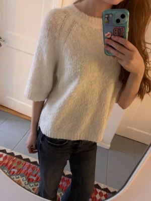 Vit stickad tröja med halvärm - Mysig vit stickad tröja med rund hals och halvärmar. Tröjan har en loose passform och är perfekt för lager-på-lager under hösten eller vintern. Materialet är mjukt och fluffigt, vilket gör den extra skön att bära. Enkel och stilren design som funkar till jeans eller kjol.