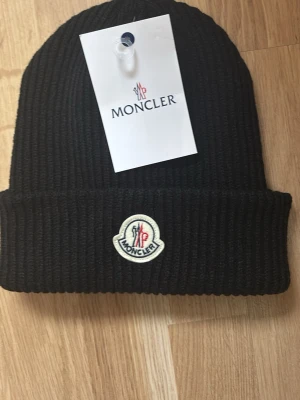 Svart ribbstickad Moncler mössa - Säljer en svart ribbstickad mössa från Moncler med uppvikt kant och klassisk Moncler-logga framtill. Mössan är i mjukt material och har en stilren look som passar till det mesta. Perfekt accessoar för kyliga dagar.