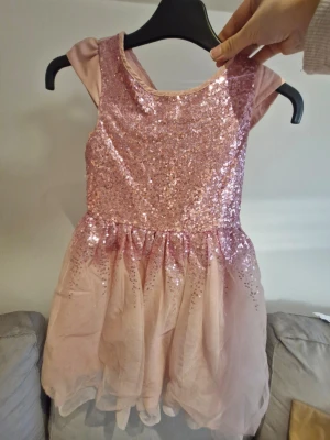 Rosa glittrig barnklänning med paljetter - Supersöt rosa barnklänning med glittriga paljetter på överdelen och ett fluffigt, skirt tyg i kjolen. Klänningen har korta ärmar och rund halsringning, samt dragkedja i ryggen för enkel påklädning. Perfekt för dig som gillar glitter och vill sticka ut.