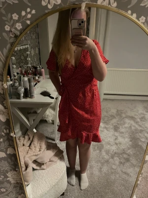Röd prickig omlottklänning - Säljer en röd kort omlottklänning med vita prickar och volangkant nedtill. Klänningen har v-ringning, korta ärmar och knyt i midjan för en snygg passform.