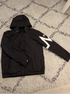 Moncler windbreaker - Svart vindjacka från Moncler med huva och dragkedja framtill. Jackan har en vit zigzag-detalj på ena ärmen och Moncler-logga. Materialet är lätt och syntetiskt, perfekt för blåsiga dagar. Fickor med dragkedja och justerbara muddar. Storleken är L men sitter lite mer som en M
