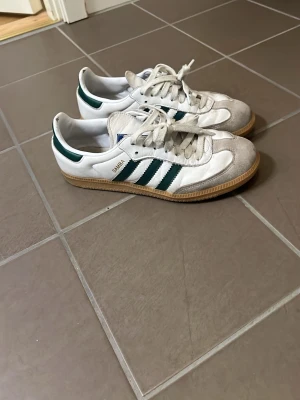 Adidas Samba sneakers vit/grön - Adidas Samba sneakers i vitt med gröna ränder och detaljer. Klassisk design med gummisula, snörning och rund tå. Ovandel i skinn och mocka, med Adidas-logga på sidan och plös. Perfekt för dig som gillar retrostil och sportig vibe.