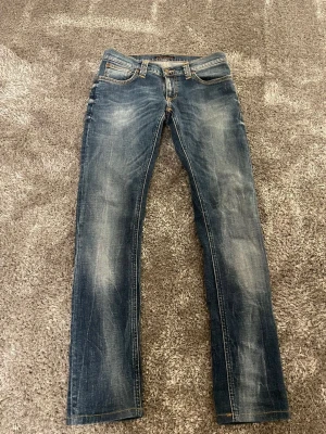 Blå jeans nudie med raka ben - Säljer ett par snygga blå jeans från nudie storlek w28 L34 med klassisk design och raka ben. Jeansen har  tvättade detaljer som ger en cool look. De har normal passform och är tillverkade i robust denim med läderpatch bak på midjan.
