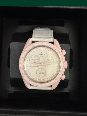 Omega swatch - Bilderna visar en Omega x Swatch Speedmaster MoonSwatch Mission to Venus klocka i ljusrosa och vitt, med biokeramisk boett och textilband. Ingen väst syns på bilderna.