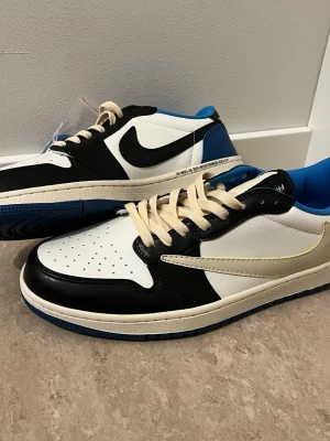 Nike Air Jordan 1 Low Travis Fragment - Nike Air Jordan 1 Low Travis Fragment med svart, vit och blå färg. Skorna har ikoniska omvända swoosh-loggor, broderade detaljer på hälen och beige snörning. Tillverkade i skinn med perforerad tå och blå yttersula. Perfekt för dig som vill sticka ut med en unik sneaker.