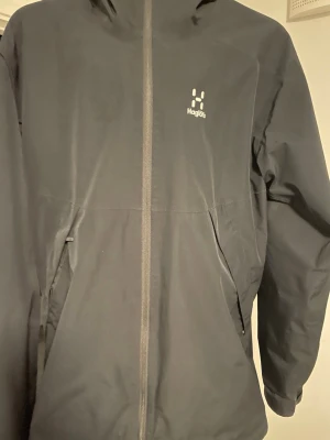 Svart vindjacka från Haglöfs - Svart vindjacka från Haglöfs med huva och hel dragkedja framtill. Jackan har två snedställda fickor med dragkedja, dragkedjan på högra fickan är sönder men annars är allt perfekt. Diskret logga på bröstet. Materialet är syntet och designen är clean och funktionell, perfekt för uteliv och äventyr.