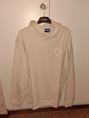 Hugo boss hoddie  - Detta är en ljusbeige, krämfärgad hoodie från HUGO i storlek L. Den har en mjuk och bekväm känsla med en normal till något avslappnad passform och en huva utan synliga snören. Framsidan är stilren med en liten rund logotyp på bröstet med texten ”Happy Hugo Love”, medan baksidan har ett större tryck med samma text i ett lekfullt och tydligt typsnitt. Helhetsintrycket är modernt och avslappnat, vilket gör hoodien lätt att bära till vardags och enkel att matcha med till exempel jeans eller mjukisby