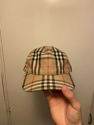 Rutig Burberry keps i beige - Säljer äkta en snygg Burberry keps med klassiskt rutig mönster i beige, svart, vitt och rött. Kepsen har justerbar rem baktill och en patch med 'Burberry London England'. Perfekt för dig som vill ha en ikonisk accessoar med street-vibe.
