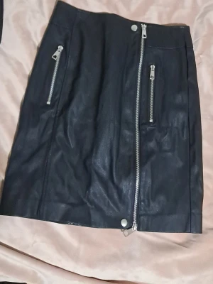 Svart skinnkjol med dragkedjor Vero Moda - Säljer en svart kort kjol från Vero Moda i skinnimitation, storlek XS. Kjolen har coola silverfärgade dragkedjor framtill och en snygg, rak passform. Perfekt för dig som gillar en edgy stil och vill ha något som sticker ut lite extra.