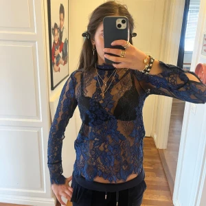 Blå spetsblus - Säljer en elegant blå spetsblus med långärmad design och hög hals. Blusen har ett transparent mönster och knytning i nacken. Perfekt för en stilfull look.