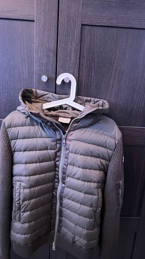 Mörk grön Moncler cardigan med huva - Säljer en mörk grön cardigan från Moncler med stickade ärmar och detaljer. Jackan har huva, två fickor med knappstängning och dragkedja framtill även en liten ficka på ärmen. Moncler-logga på vänster ärm har självklart NFC Scann, och ribbade muddar. Perfekt för dig som vill ha en snygg och varm jacka med snygga detaljer.