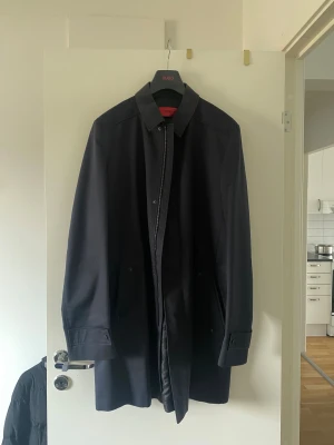 Svart rock från Hugo Boss - Snygg svart rock från Hugo Boss med klassisk krage och diskreta knappar. Insidan har coola röda detaljer och ficka med knapp. Materialet är slätt och känns lyxigt, perfekt för dig som vill ha en stilren look. Ärmarna har justerbara remmar med knapp.