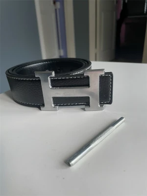 H bälte  - Snyggt svart skärp med ett stort H-format spänne i silverfärgad metall. Skärpet är gjort av präglat skinn  vita sömmar längs kanterna för en cool kontrast. Perfekt accessoar för att lyfta din outfit.