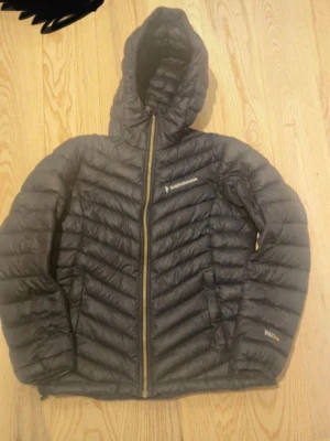Svart dunjacka Peak Performance M - Svart dunjacka från Peak Performance med huva och snygga quiltade detaljer. Jackan har dragkedja framtill och logga på bröstet. Perfekt för kalla dagar och har en modern, sportig look. Passar dig som gillar stil och funktion. Original pris 3200kr