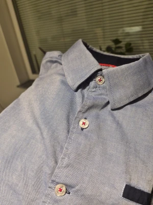 Blå skjorta från Savvy Citizen M - Snygg ljusblå skjorta från Savvy Citizen i bomull, med klassisk krage och långa ärmar. Knapparna har röd tråd och detaljer i mörkblått vid bröstfickan och manschetterna. Perfekt för dig som gillar stilrena och moderna plagg.