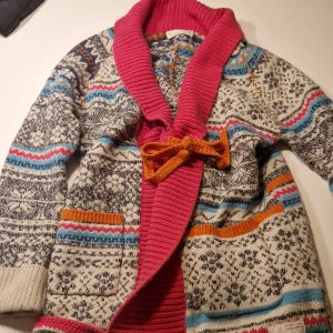 Mönstrad  odd molly kofta med rosa krage - Säljer en unik och färgglad odd molly  kofta med mönster i svart, vit, blå och orange. Koftan har en bred, ribbad rosa krage och ett orange knytband framtill. Två fickor med orange kant och långa ärmar. Perfekt för dig som gillar stickade plagg med mycket färg.